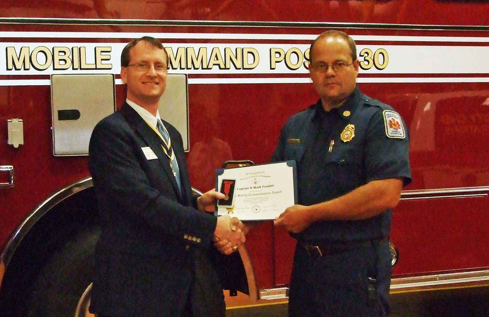 Mark Feaster receives the Fairfax Resolves Fire Safety Commendation from Chapter President Darrin Schmidt. The station's mobile command post vehicle serves as a backdrop.